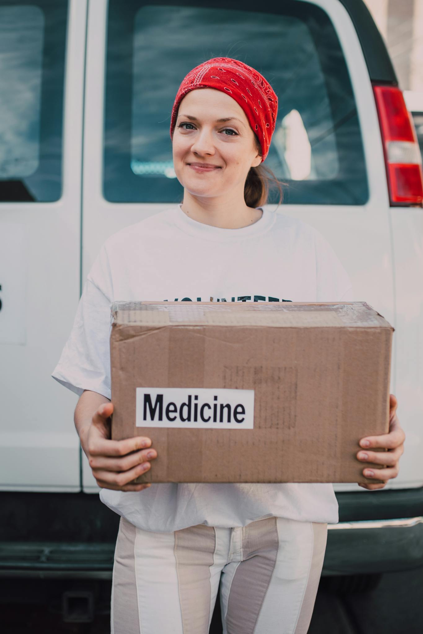 A smiling female volunteer holding a donation box labeled 'Medicine' in front of a van.